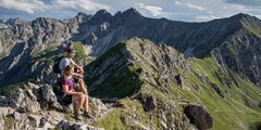 A couple rests on a rocky mountain summit, enjoying the panoramic view of a majestic mountain range. © Sport Kessler GmbH