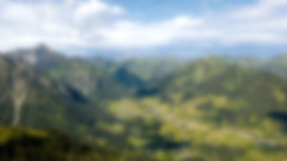 A panoramic view of a valley nestled within the Alps, showcasing a charming village surrounded by lush green hills and towering mountains under a partly cloudy sky. © Sport Kessler GmbH
